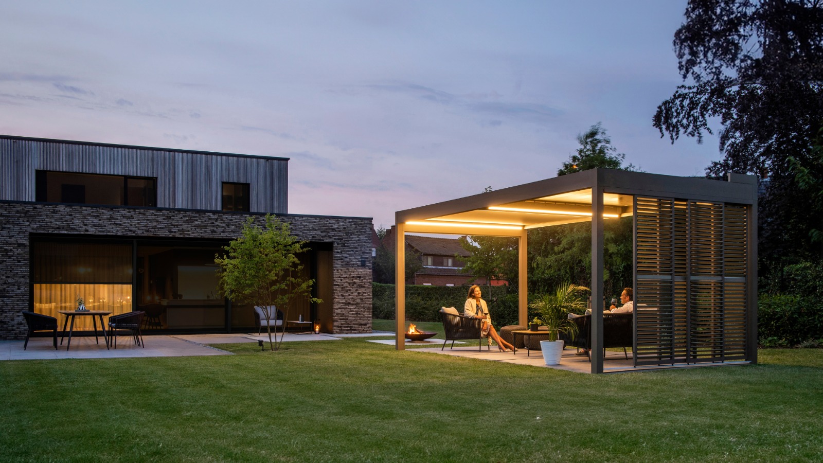Pergolă Hogarti iluminată seara, cu zonă lounge, lângă o casă modernă